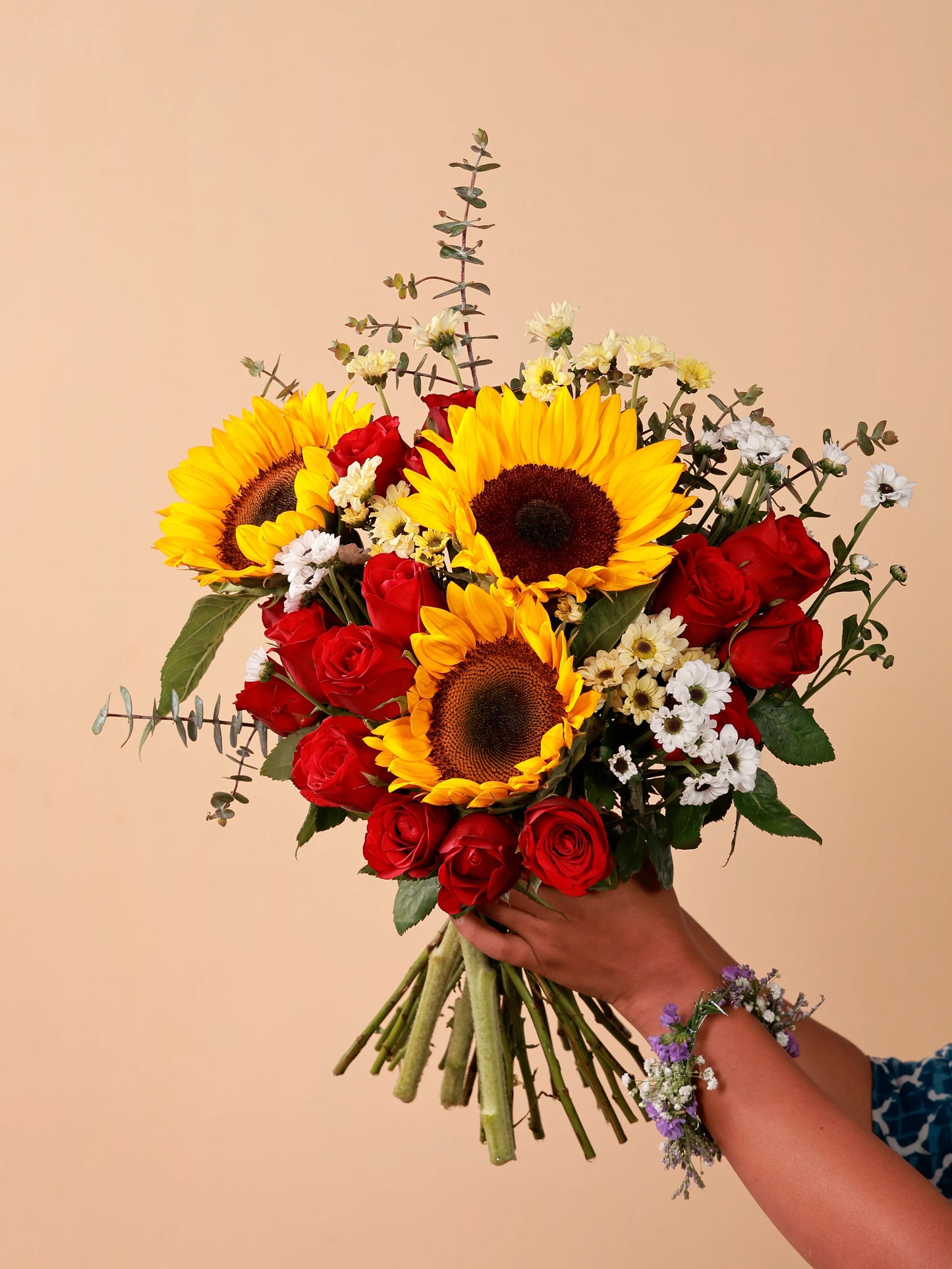 A hand holding a bouquet of sunflowers, red roses, and white santinis